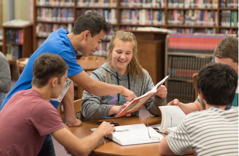A group of students in a library reviewing a textbook together.